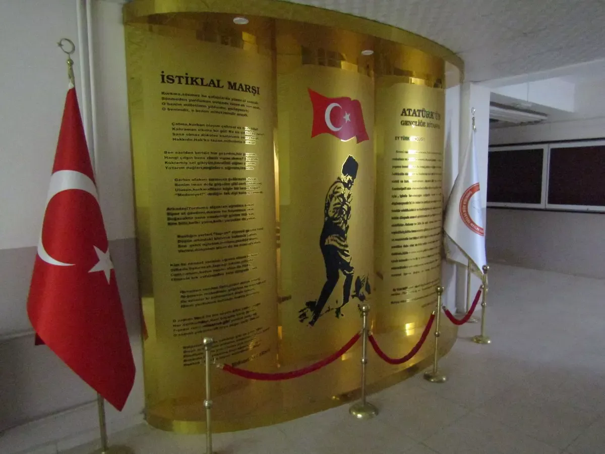 Bir okulun koridorunda iki Türk bayrağı arasında, İstiklal Marşı, Atatürk silueti ve Gençliğe Hitabe metninin yer aldığı üç bölmeli altın renkli pano