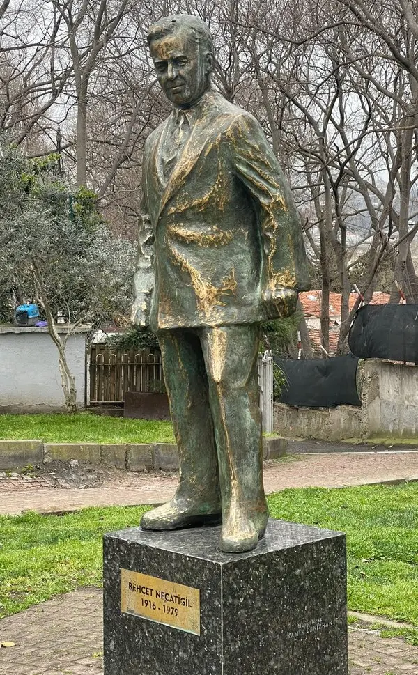 Bir parkta siyah taş kaide üzerinde duran, takım elbiseli Behçet Necatigil'i ayakta gösteren bronz heykel; kaidede şairin adı ve 1916–1979 yazılı