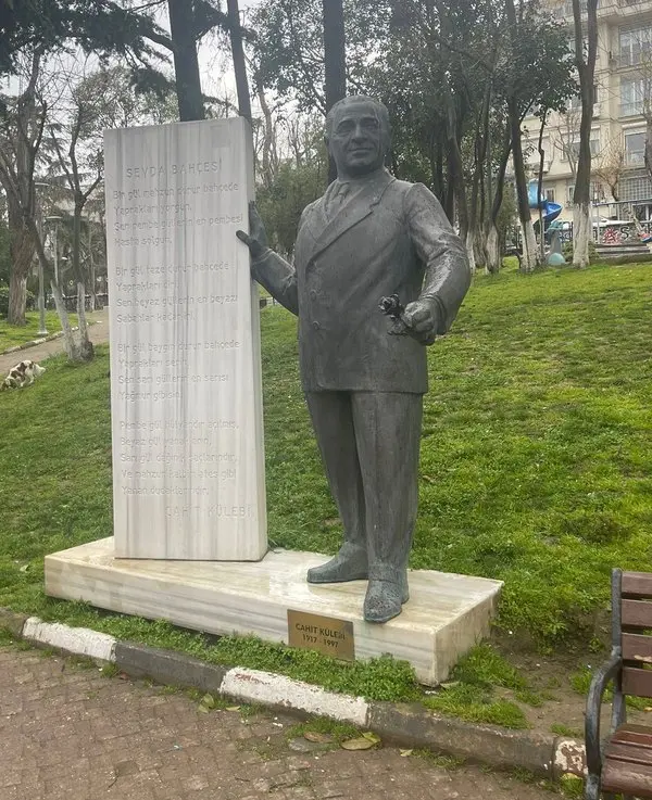 Çimenlik bir parkta, üzerinde şiir yazılı beyaz mermer bir anıtın yanında duran, takım elbiseli Cahit Külebi'nin koyu renkli heykeli