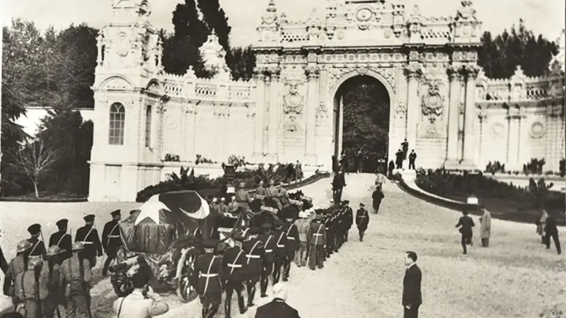 Atatürk'ün cenaze alayının Dolmabahçe Sarayı'nın süslü kapısından çıkışı: bayrağa sarılı tabutu taşıyan top arabası ve iki yanında yürüyen askerler, siyah-beyaz fotoğraf