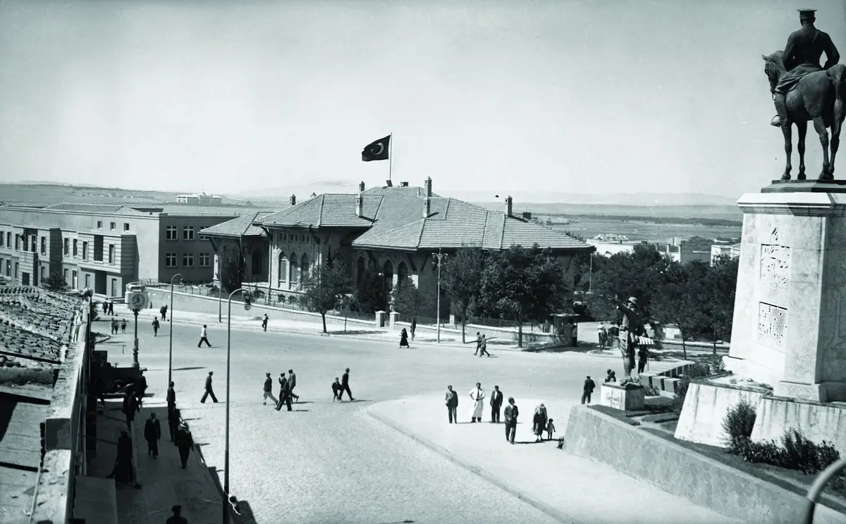 1930'larda Ankara Ulus Meydanı: sağda atlı Zafer Anıtı, arkada çatısında bayrak dalgalanan ilk Türkiye Büyük Millet Meclisi binası ve meydanda yürüyen insanlar
