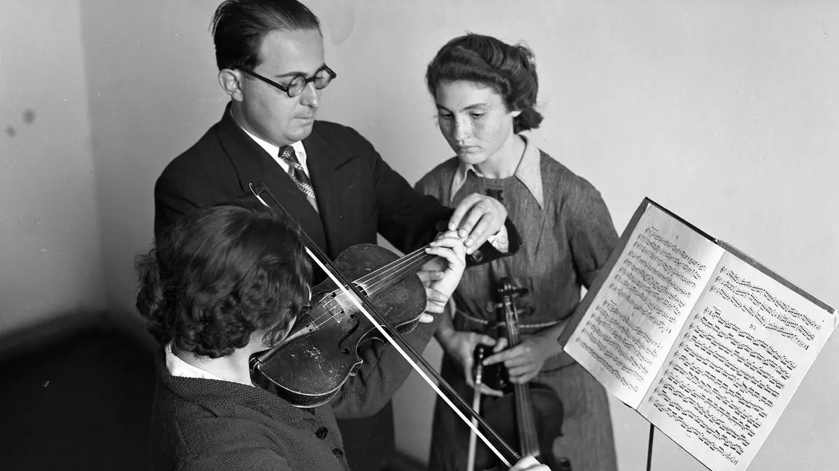 1930'larda Ankara Musiki Muallim Mektebi'nde gözlüklü öğretmenin keman çalan iki kız öğrenciye nota sehpası başında ders verişi, siyah beyaz