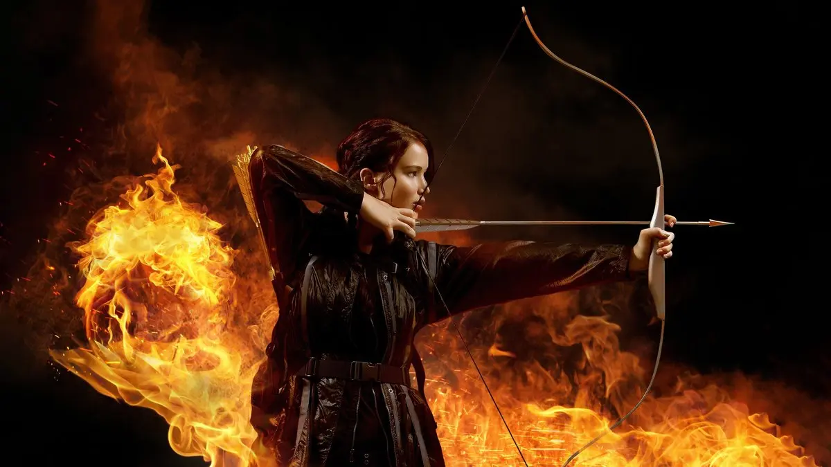 Açlık Oyunları filminden tanıtım görseli: siyah deri ceketli Katniss Everdeen, alevlerin önünde yayını gererek oku yana doğru nişan alıyor