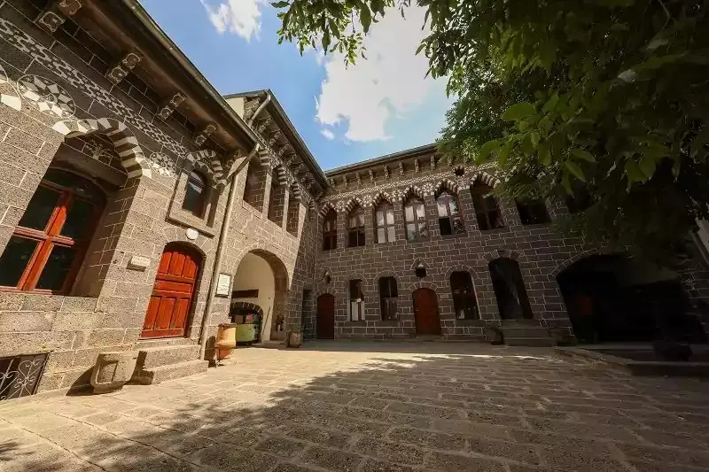 Diyarbakır Sur’daki Ahmet Arif Edebiyat Müzesi’nin bazalt taşından avlusu; kırmızı ahşap kapılar, kemerli pencereler ve avluya gölge düşüren büyük bir ağaç