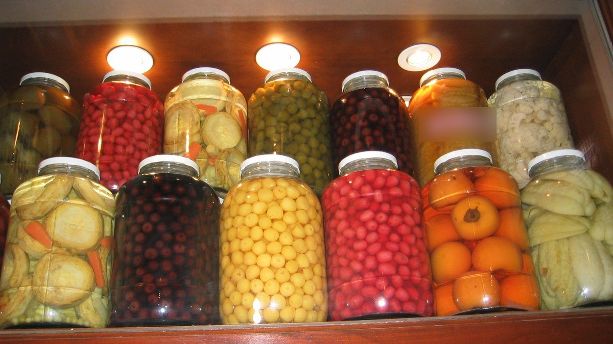 Işıklı bir rafta yan yana dizilmiş, salatalık, biber, zeytin ve kızılcık dolu renkli turşu kavanozları