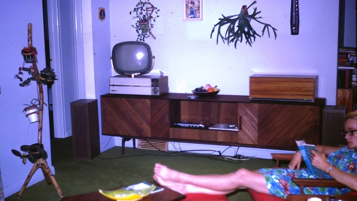 1960'lardan kalma bir oturma odasında ahşap konsolun üzerinde duran küçük tüplü televizyon ve kitap okuyan bir kadın