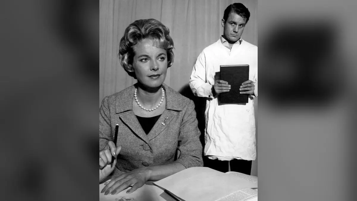 The Two Worlds of Charlie Gordon (1961) televizyon uyarlamasından siyah beyaz bir kare: masada kalemle oturan öğretmen Alice rolünde Mona Freeman, arkasında beyaz önlüğüyle elinde kitap tutan Charlie Gordon rolünde Cliff Robertson