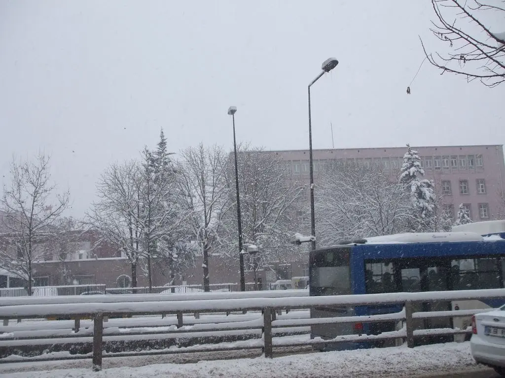 Kar yağarken Ankara Bakanlıklar’da bir cadde; karla kaplı ağaçlar ve korkuluklar, arkada pembe cepheli bir kamu binası ve mavi bir belediye otobüsü