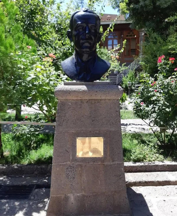 Ağaçlı bir bahçede taş kaide üzerinde duran, Cahit Sıtkı Tarancı'yı gösteren koyu renkli bronz büst