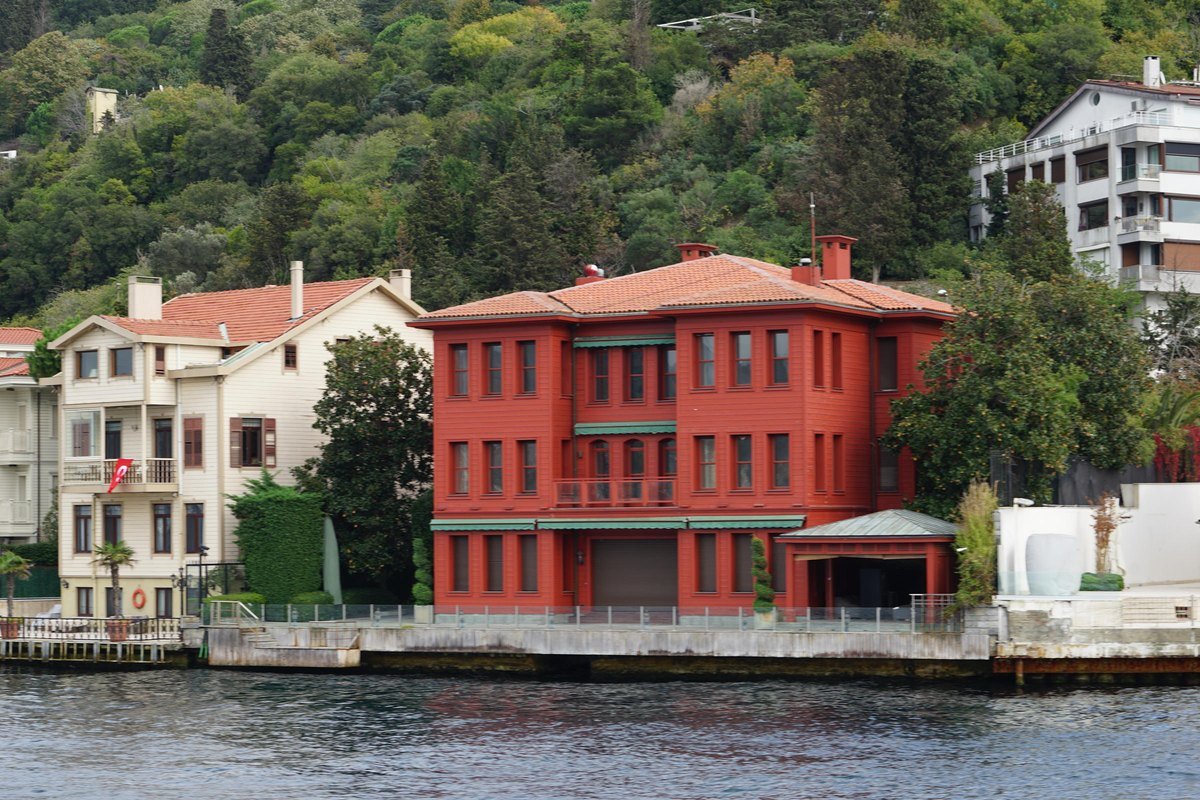 Boğaz kıyısında, ormanlık yamacın önünde denize sıfır duran kırmızı boyalı büyük bir yalı ve yanındaki beyaz evler