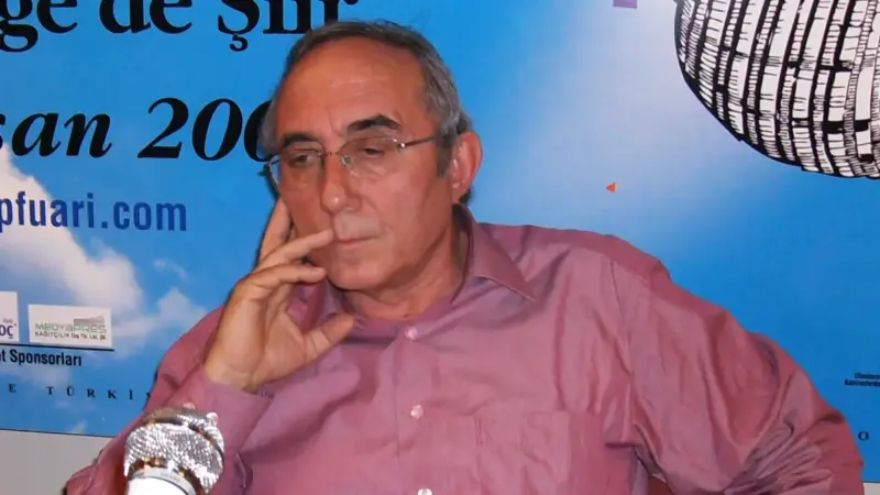 Ataol Behramoğlu, pembe gömlek ve gözlükle elini yüzüne dayamış, önünde mikrofonla 13. İzmir Kitap Fuarı’ndaki söyleşide