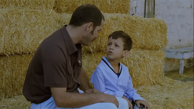 Babam ve Oğlum filminden sahne: saman balyalarının önünde oturan Sadık, yanındaki beyaz denizci yakalı küçük oğlu Deniz’e eğilmiş konuşuyor