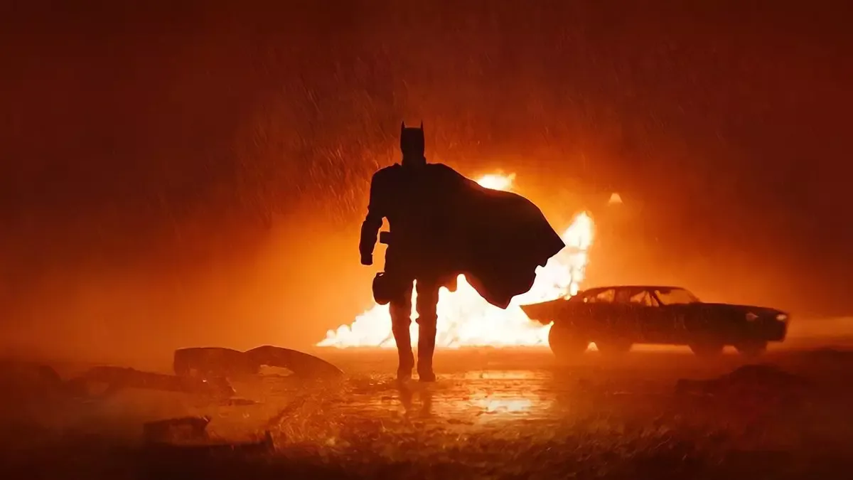 The Batman filminden sahne: yağmurlu gecede arkasında alevler yükselen bir arabanın önünden pelerini dalgalanarak yürüyen Batman rolündeki Robert Pattinson