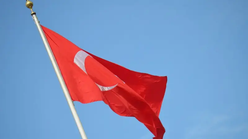 Mavi gökyüzü önünde, altın topuzlu beyaz bir direkte rüzgârla dalgalanan ay yıldızlı al Türk bayrağı