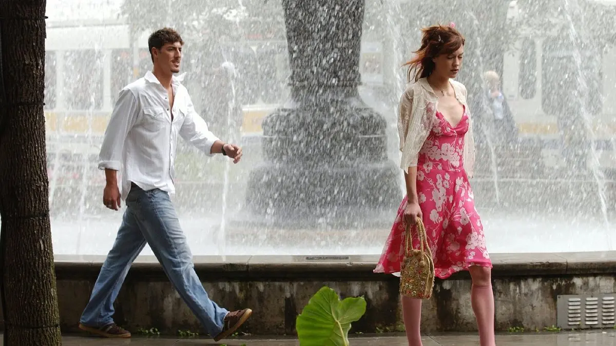 Benimle Ol (Lie with Me, 2005) filminden bir kare: yaz günü bir fıskiyenin önünden geçen, pembe çiçekli elbiseli ve elinde hasır çanta tutan Lauren Lee Smith’in arkasından beyaz gömlekli Eric Balfour gülümseyerek yürüyor