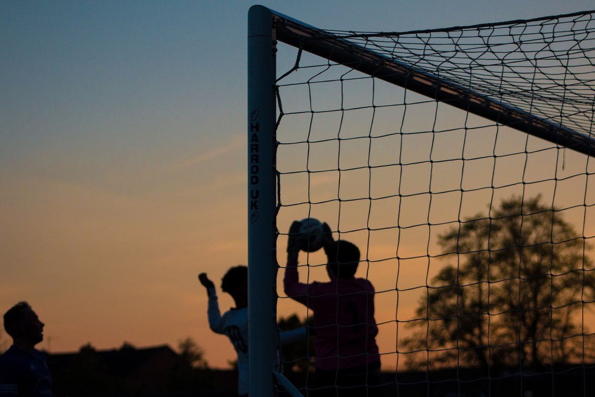 Gün batımında futbol kalesinin önünde topu havada yakalayan kalecinin ve oyuncuların siluetleri