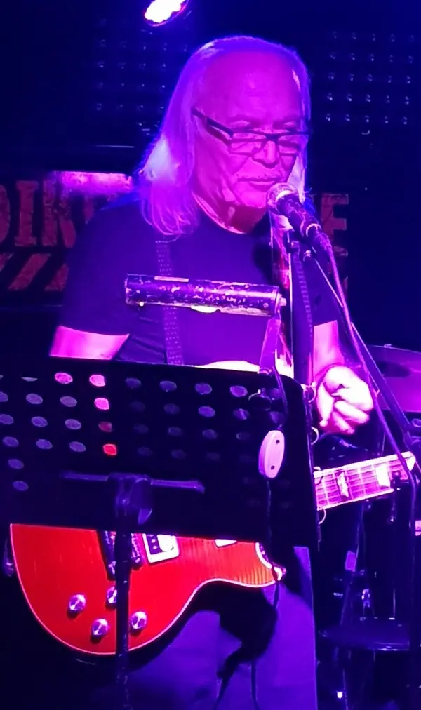 Uzun beyaz saçlı ve gözlüklü Cahit Berkay sahnede mikrofon önünde, kırmızı elektro gitarını çalarken