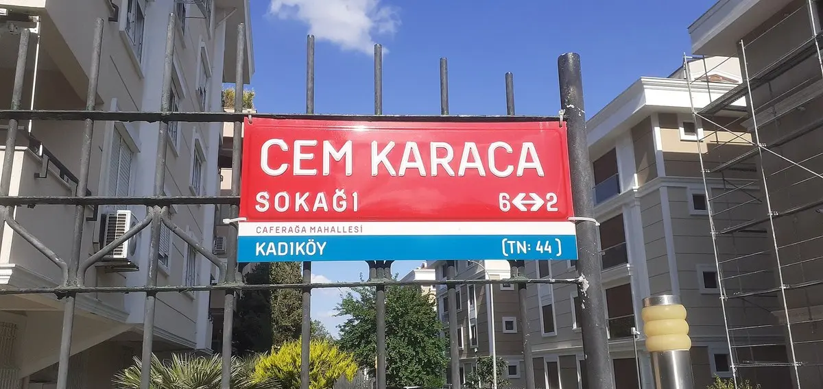 Kadıköy’de bir demir parmaklık üzerine asılı kırmızı-beyaz “Cem Karaca Sokağı, Caferağa Mahallesi, Kadıköy” tabelası, arkada apartmanlar ve mavi gökyüzü