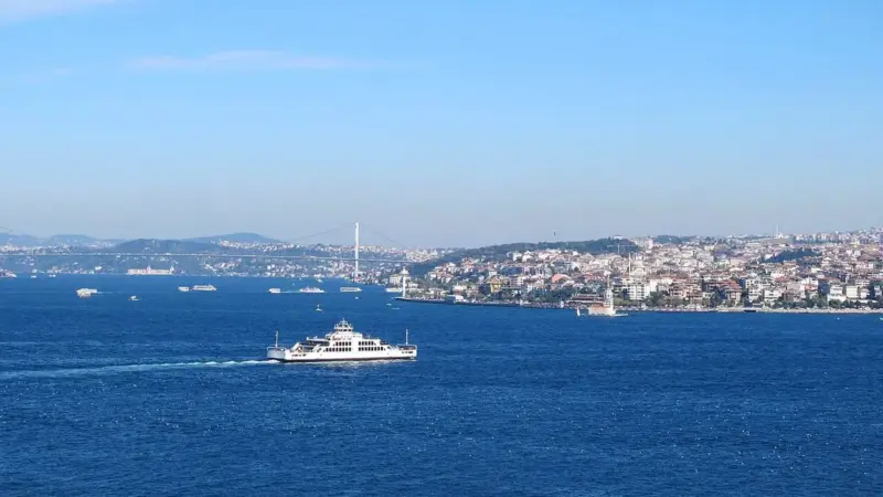 Masmavi İstanbul Boğazı’nda ilerleyen beyaz bir feribot; arka planda Boğaziçi Köprüsü, Kız Kulesi ve Üsküdar kıyısı
