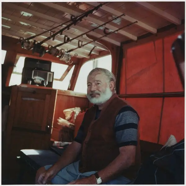 Beyaz sakallı Ernest Hemingway'in teknesi Pilar'ın kırmızı ahşap kamarasında, çizgili tişört ve yelekle oturup kameraya gülümsediği renkli fotoğraf