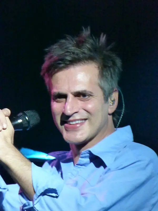 Açık mavi gömlekli Teoman'ın karanlık bir sahnede elinde mikrofonla gülümseyerek şarkı söylediği konser fotoğrafı
