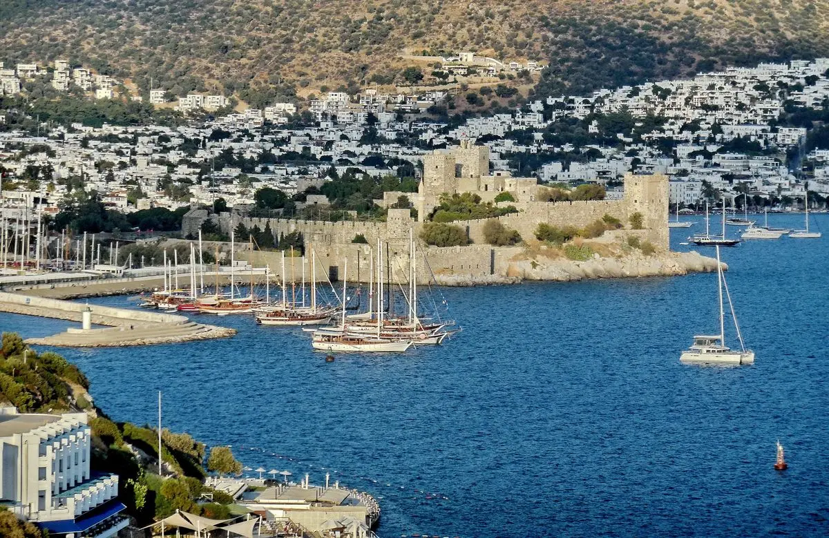 Bodrum Kalesi'nin mavi koya uzanan surları, önünde demirli yelkenliler ve arkada beyaz evlerle kaplı Bodrum yamaçları