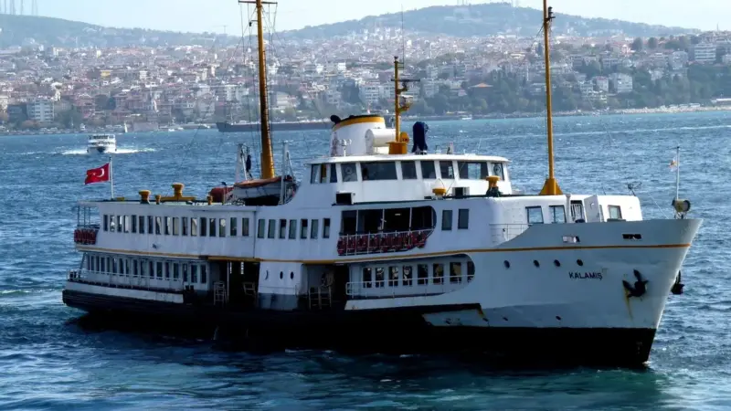 İstanbul Boğazı'nın mavi sularında ilerleyen beyaz gövdeli Kalamış vapuru; arkada kıyı boyunca sıralanan evler ve yeşil sırtlar