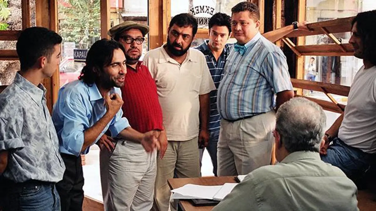 Ekmek Teknesi dizisinden bir mahalle kahvesinde masanın başında oturan adamın çevresinde ayakta toplanmış gençler ve mahalle sakinleri