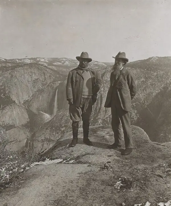 Siyah beyaz fotoğraf: Yosemite Vadisi’nde Glacier Point’in kayalık zirvesinde yan yana duran şapkalı Theodore Roosevelt ve uzun sakallı John Muir, arkalarında karlı dağlar, şelale ve derin vadi