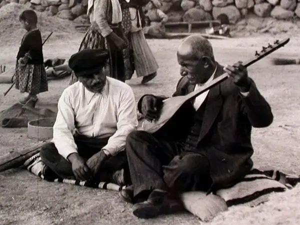 Âşık Veysel’in yere serili bir kilimin üzerinde bağdaş kurup bağlama çaldığı, yanında oturan bir gencin onu dinlediği siyah-beyaz fotoğraf