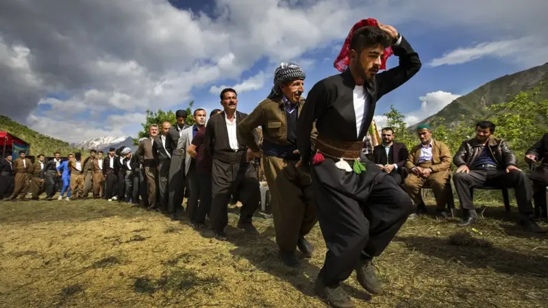 Anadolu’da açık havada yapılan bir düğünde başında kırmızı mendil tutan genç bir adamın önderliğinde yan yana dizilip halay çeken, geleneksel kıyafetli erkekler; arkada dağlar ve bulutlu gökyüzü