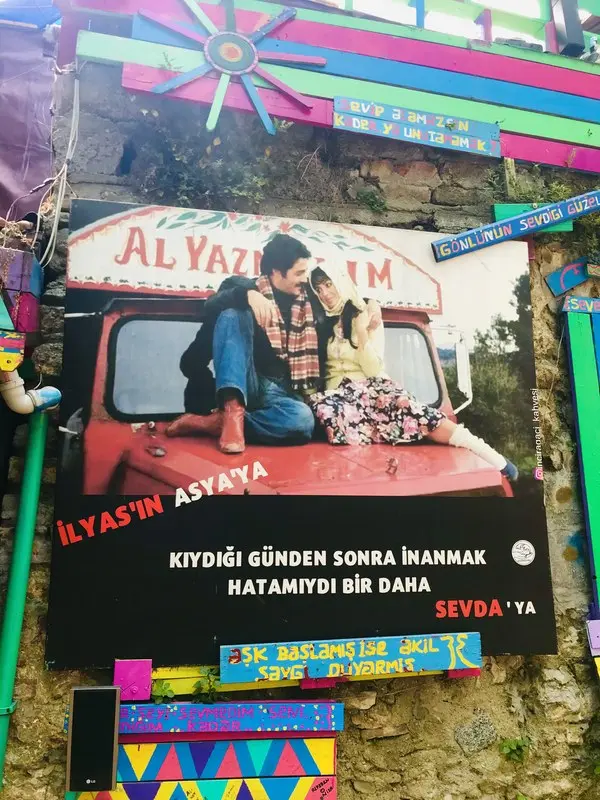 Balat’ta renkli bir duvara asılmış Selvi Boylum Al Yazmalım afişi: kırmızı kamyonun kaputunda oturan İlyas ve Asya