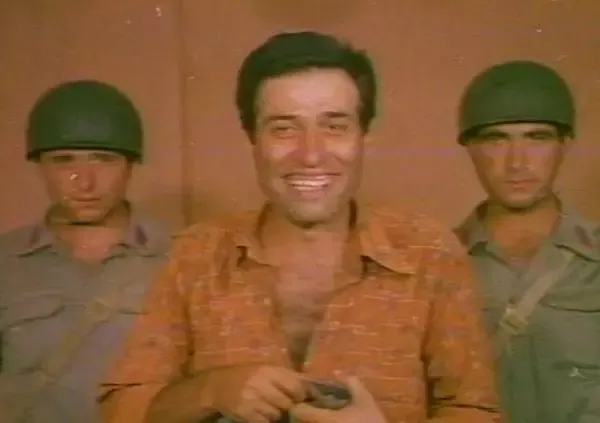 Kibar Feyzo filminden bir kare: turuncu gömlekli Kemal Sunal gülümserken iki yanında miğferli askerler