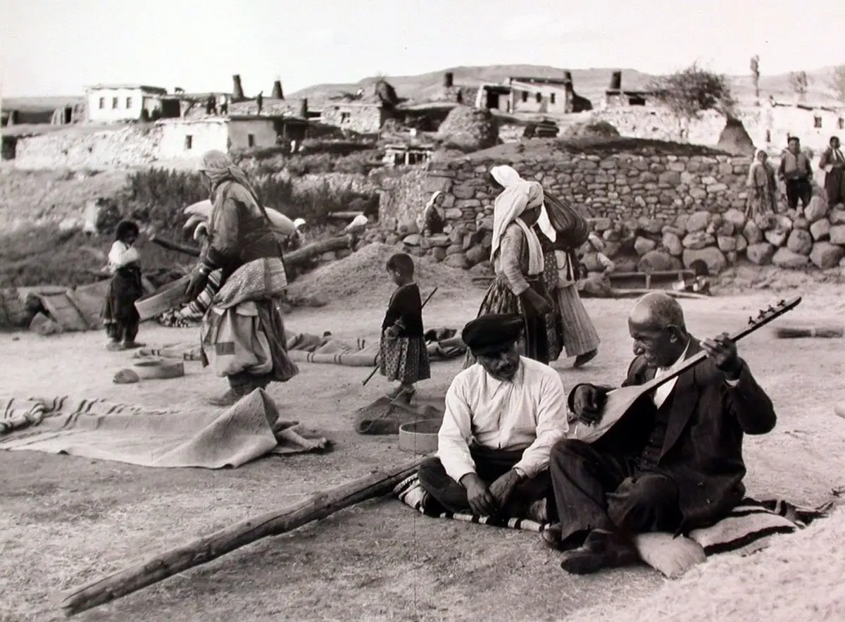 Âşık Veysel köy evlerinin önünde yere oturmuş bağlama çalarken, çevresinde köylüler; siyah beyaz fotoğraf