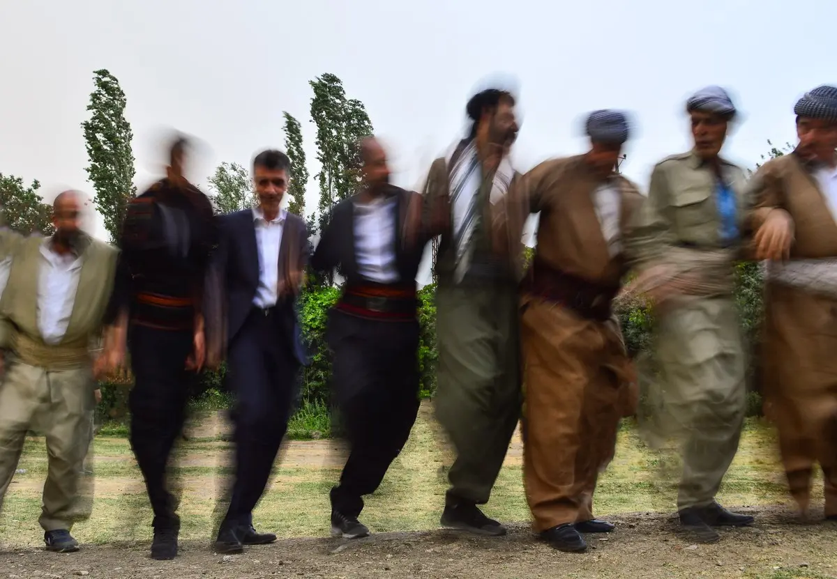 Açık havada yan yana kol kola girmiş, halay çeken bir grup erkek; bazıları yöresel kıyafetli