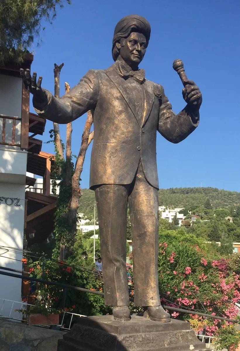 Bodrum’da mavi gökyüzü altında, elini kaldırmış ve mikrofon tutan Zeki Müren’in bronz heykeli