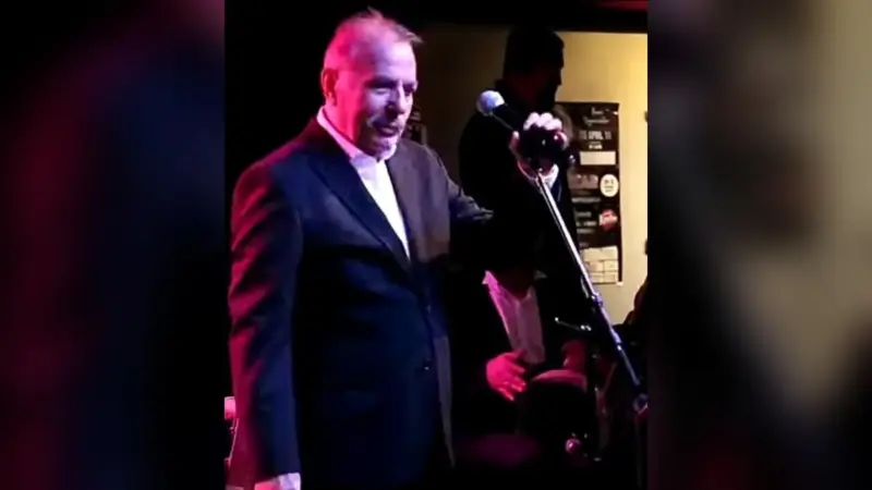Ferdi Tayfur’un 2019’daki Münih konserinde koyu takım elbise ve beyaz gömlekle sahnede, mikrofon sehpasının başında şarkı söylerken çekilmiş fotoğrafı