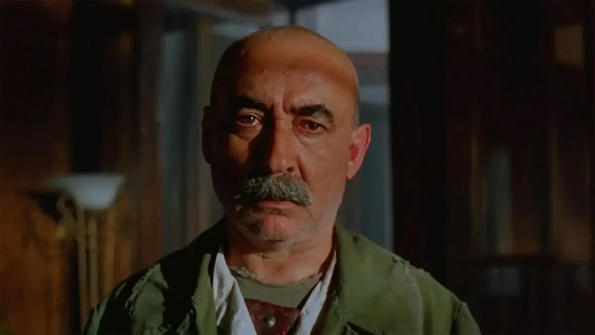 Eşkıya filminden, loş bir otel odasında haki ceketli, kır bıyıklı Baran rolündeki Şener Şen'in ciddi bakışlı yakın planı