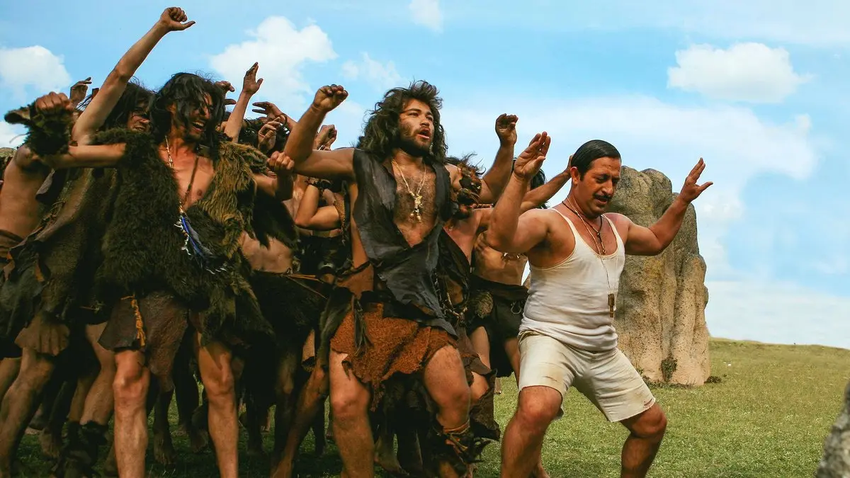 A.R.O.G filminden, yeşil bir düzlükte dikili taşların önünde kollarını havaya kaldırarak coşkuyla dans eden hayvan postlu mağara insanları ve beyaz atletli Arif
