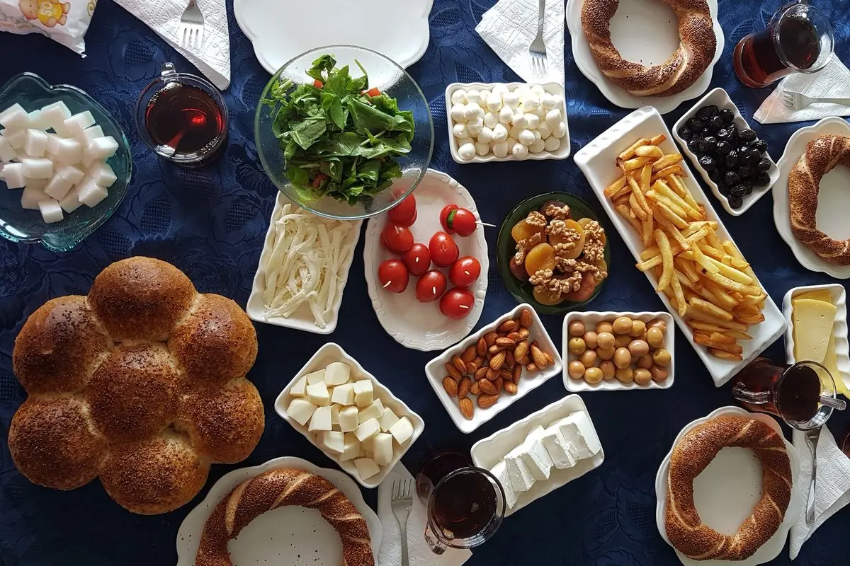 Lacivert örtülü masada kurulmuş Türk kahvaltısı; simitler, susamlı çörek, beyaz peynir, kaşar, zeytin, domates, roka, kuruyemiş, patates kızartması ve ince belli bardaklarda çay