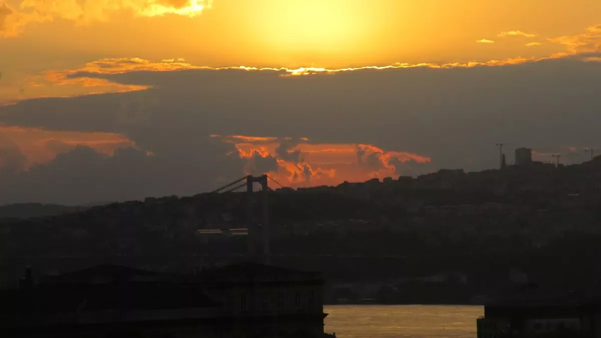 İstanbul Boğazı’nda gün doğumu; turuncu ve altın renkli gökyüzünde bulutların arkasından yükselen güneş, siluet hâlinde Boğaz köprüsü, Anadolu yakasının tepeleri ve ışıltılı deniz