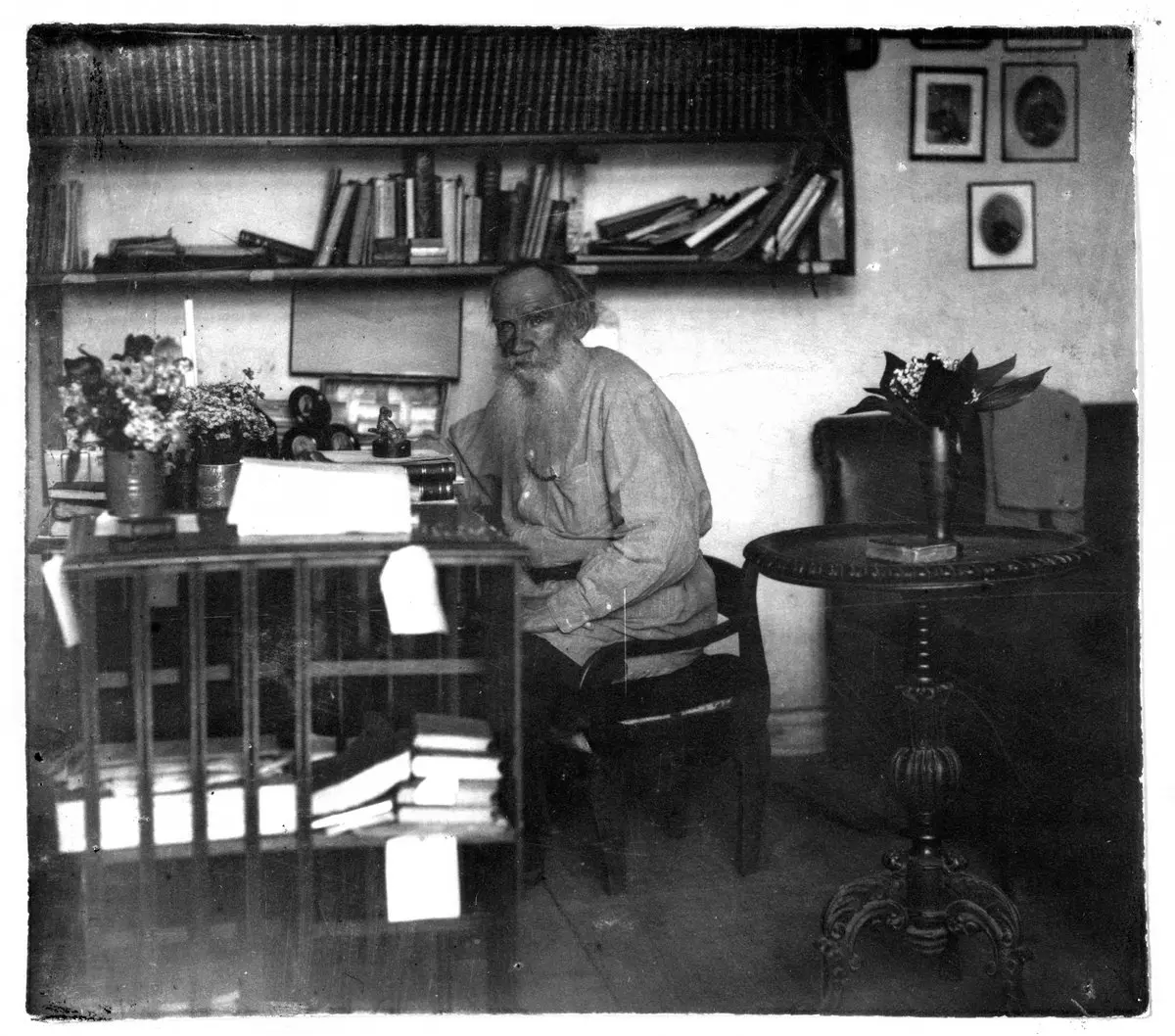 Lev Tolstoy, kitap rafları ve yazı masasıyla çevrili çalışma odasında sandalyede oturuyor; 1908 tarihli siyah beyaz fotoğraf
