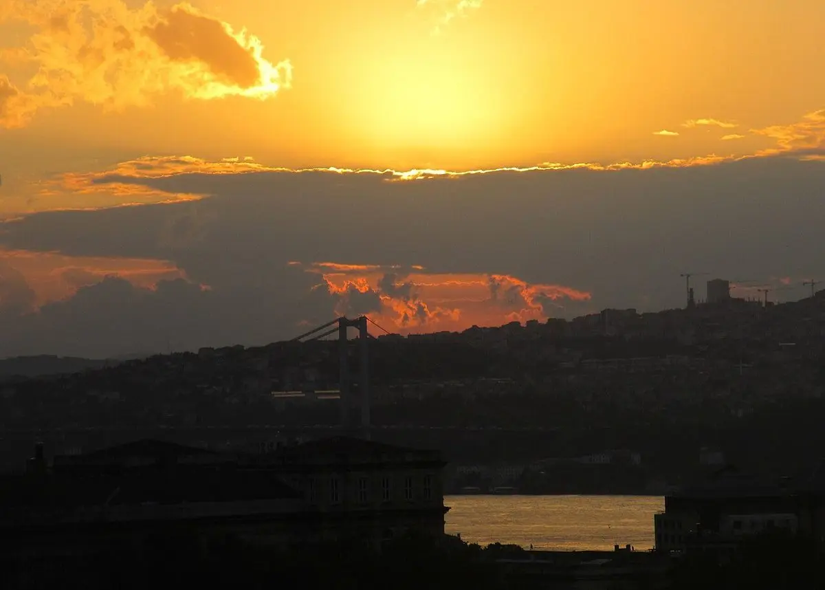 İstanbul Boğazı’nda gün doğumu: Turuncu bir gökyüzünün altında karşı kıyının karartısı ve Boğaz köprüsünün silueti, ön planda sakin su