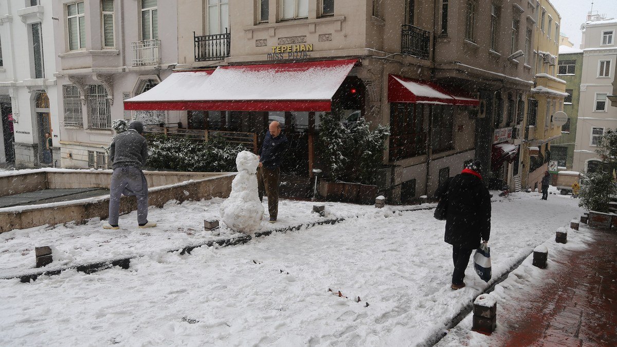 Beyoğlu Asmalımescit'te karla kaplı bir sokakta kardan adam yapan insanlar ve kırmızı tenteli eski bir bina