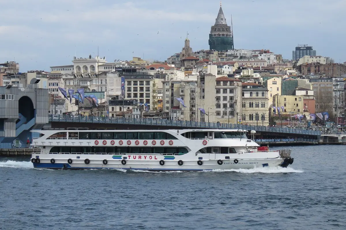 Galata Köprüsü önünden geçen beyaz bir yolcu vapuru, arkada yamaçtaki binalar ve Galata Kulesi