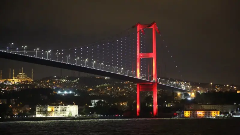 Gece İstanbul’da kırmızı ışıklarla aydınlatılmış Boğaziçi Köprüsü ve arkasında ışıl ışıl kıyı