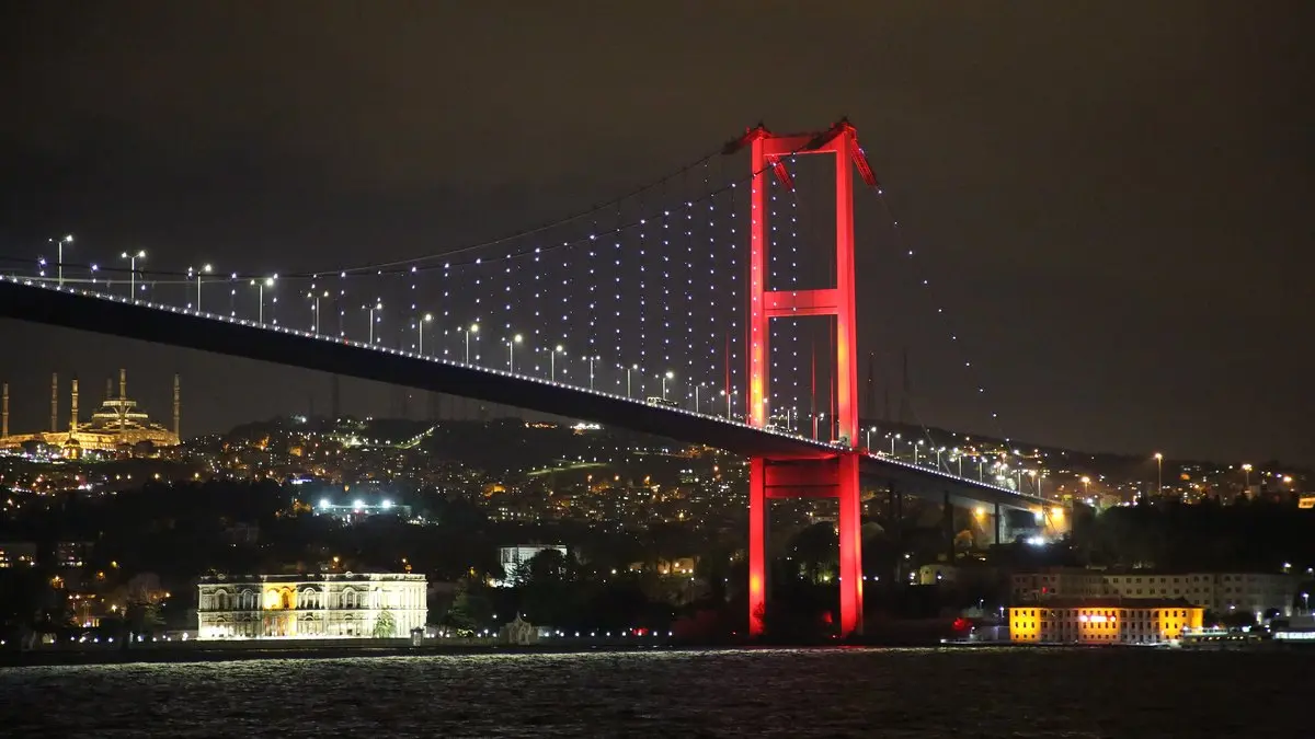 Gece İstanbul’da kırmızı ışıklarla aydınlatılmış Boğaziçi Köprüsü ve arkasında ışıl ışıl kıyı