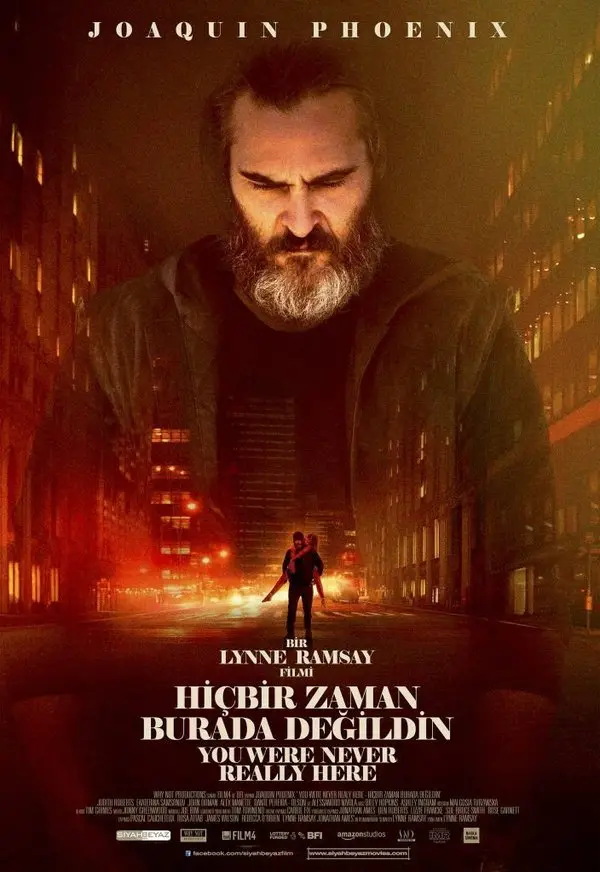 Hiçbir Zaman Burada Değildin filminin Türkçe afişi: sakallı Joaquin Phoenix'in yüzü kırmızı turuncu şehir ışıklarının üzerine bindirilmiş; altta yürüyen küçük bir insan silueti