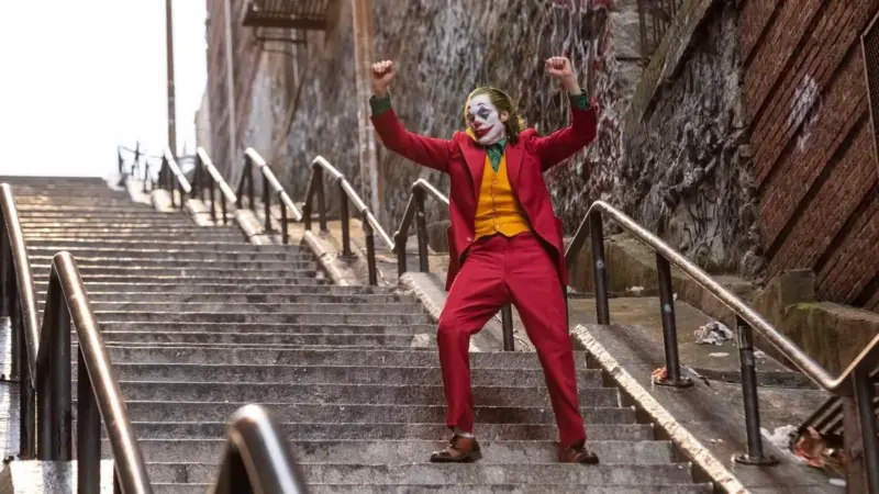 Joker filminden tanıtım karesi: kırmızı takım elbiseli, hardal yelekli ve palyaço makyajlı Arthur Fleck, grafitili tuğla duvarlar arasındaki uzun taş merdivende kollarını havaya kaldırmış dans ediyor