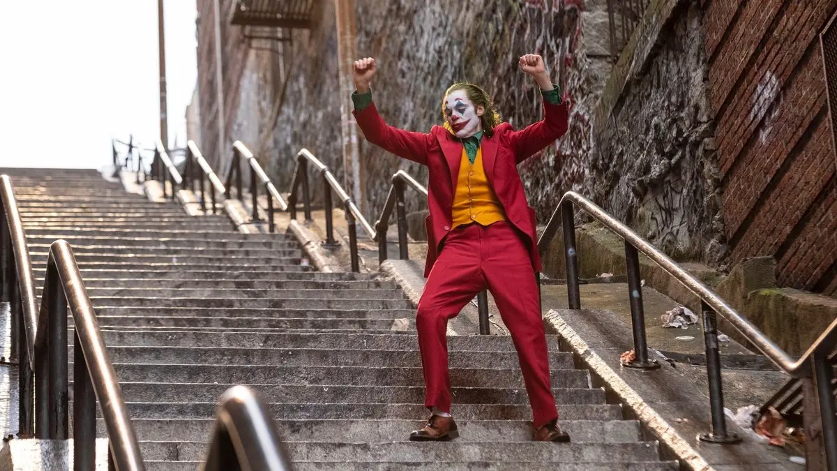 Joker filminden tanıtım karesi: kırmızı takım elbiseli, hardal yelekli ve palyaço makyajlı Arthur Fleck, grafitili tuğla duvarlar arasındaki uzun taş merdivende kollarını havaya kaldırmış dans ediyor